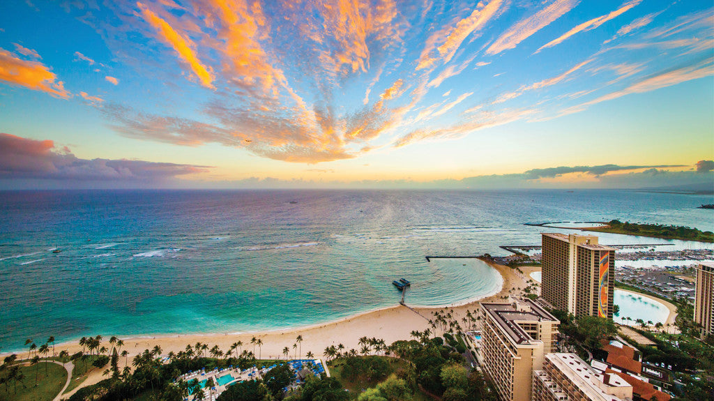 A panoramic view of the Hilton Hawaiian Village and the ocean during sunset hour, casting a warm golden glow across the landscape.